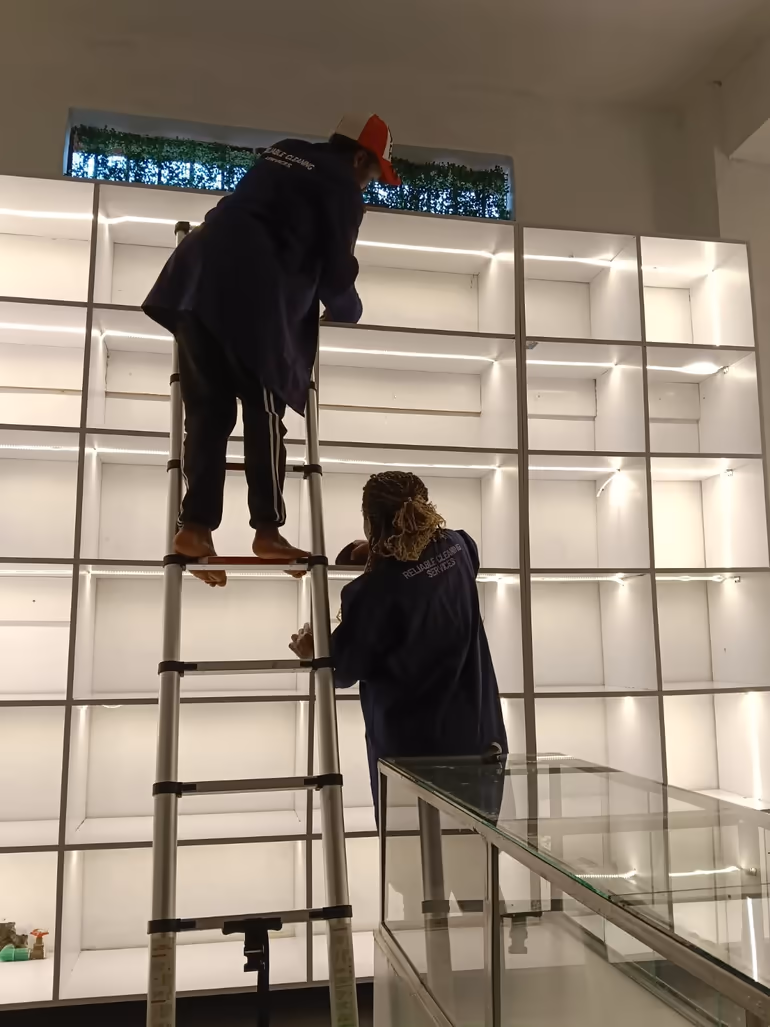 Two Shine On cleaners on a ladder cleaning tall backlit retail display shelves in Nairobi